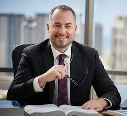 Smiling man in a suit holding glasses, sitting with an open book in an office setting.