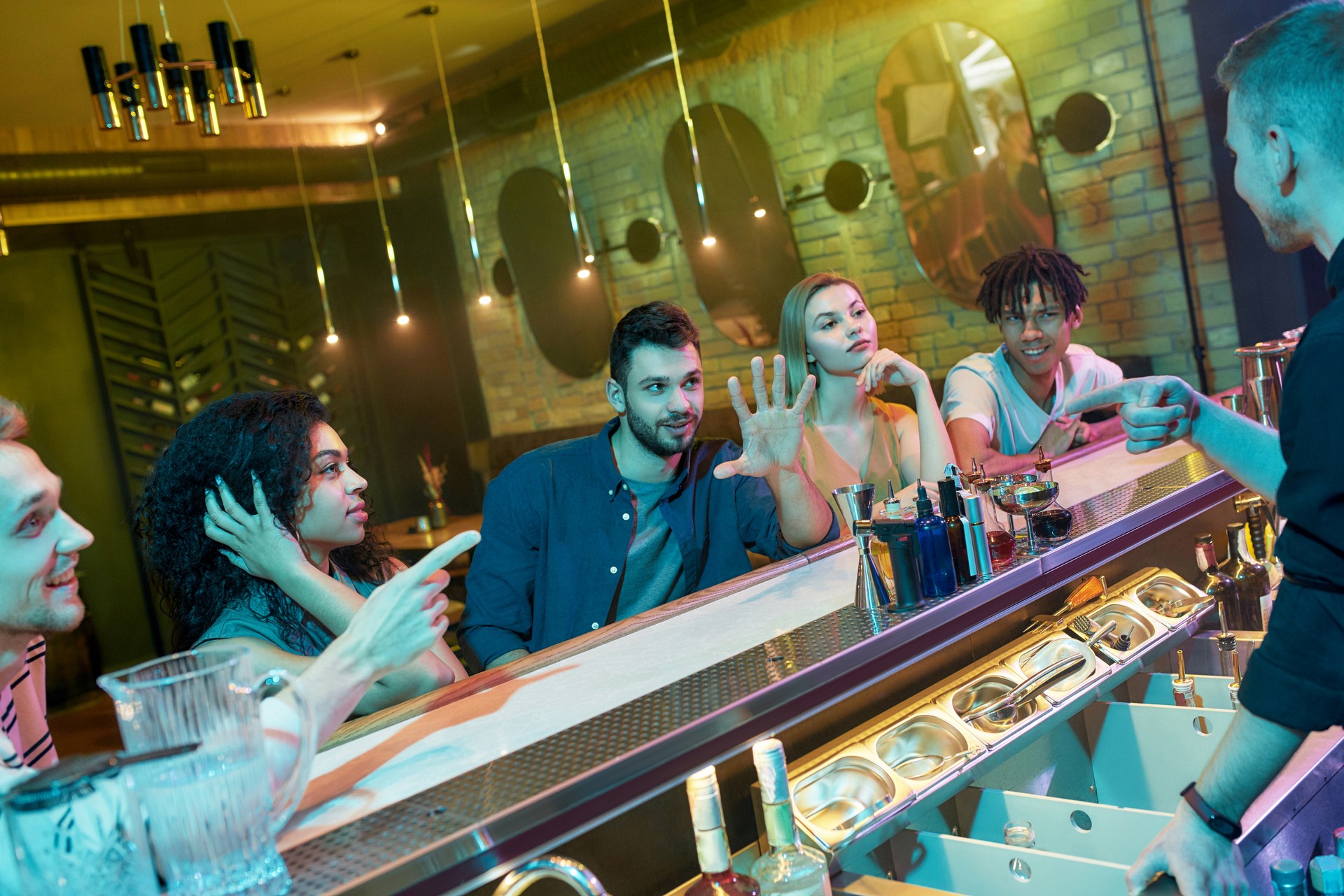 Young man ordering drinks for his friends, talking to male bartender. Group of young adults, men and women having fun, waiting for cocktails at the bar counter at the night club